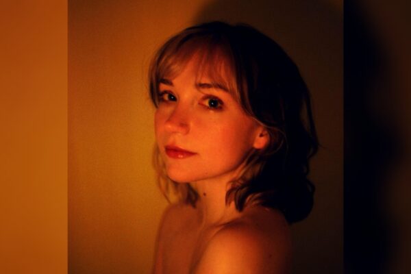 The album cover for musical artist Flower Face's "Girl Prometheus," featuring a young white woman softly smiling while glancing over her shoulder on a dark orange and black background. Her face is dimly lit.