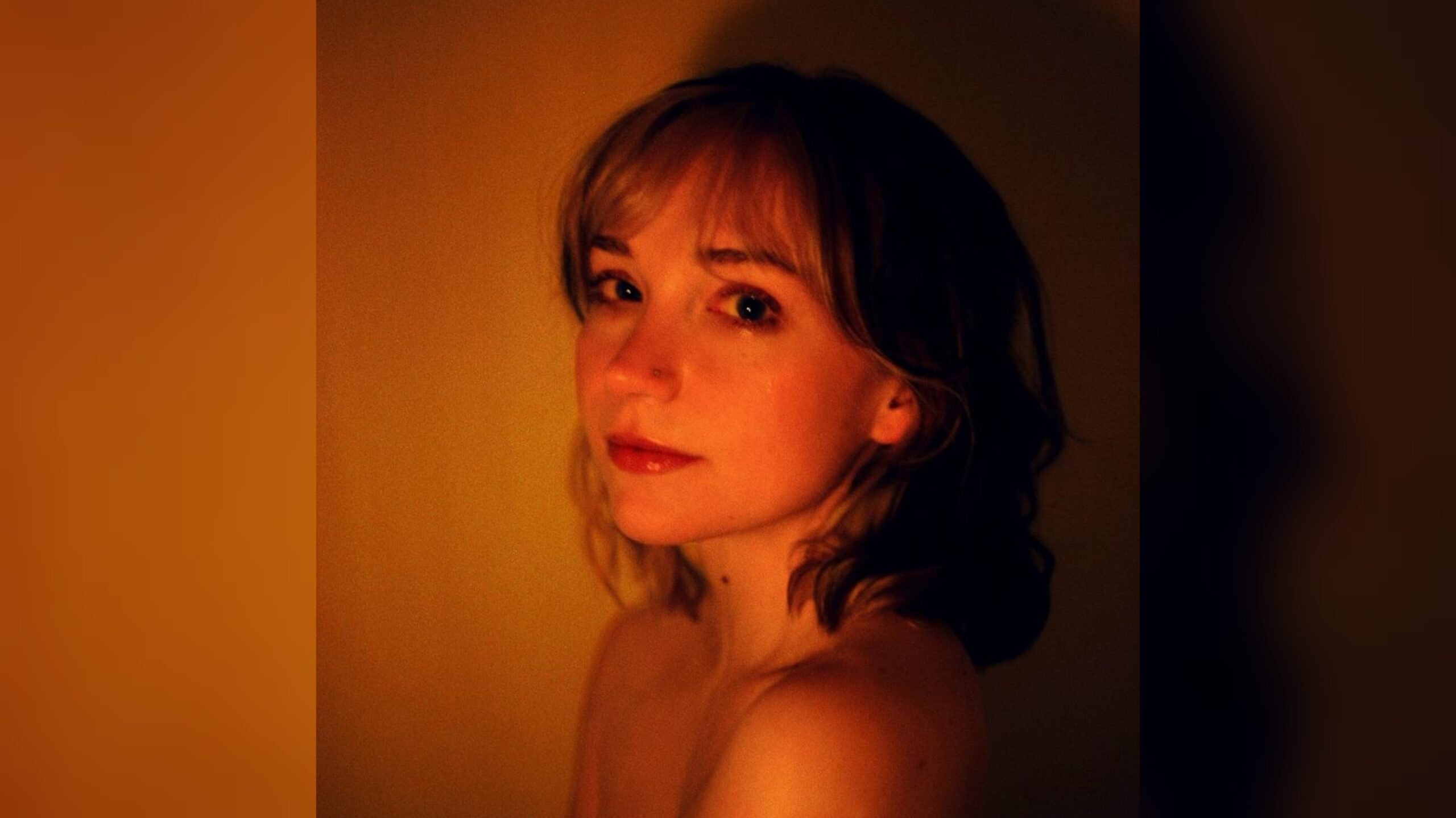 The album cover for musical artist Flower Face's "Girl Prometheus," featuring a young white woman softly smiling while glancing over her shoulder on a dark orange and black background. Her face is dimly lit.