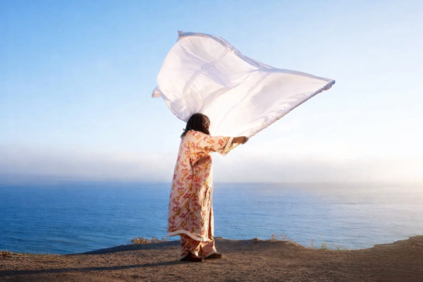 Elmiene’s ‘sounds for someone’: A Debut That Finds Power in Simplicity — Album Review Elmiene stands on a cliff by the ocean holding a flowing white fabric against a clear sky