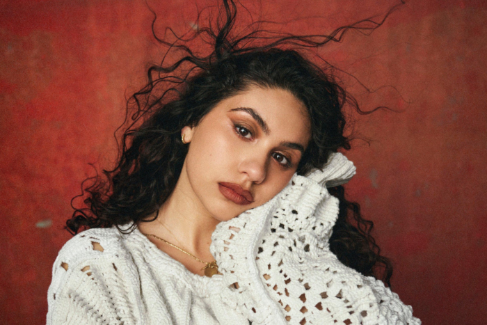 Alessia Cara in a white knit sweater posing against a textured red background