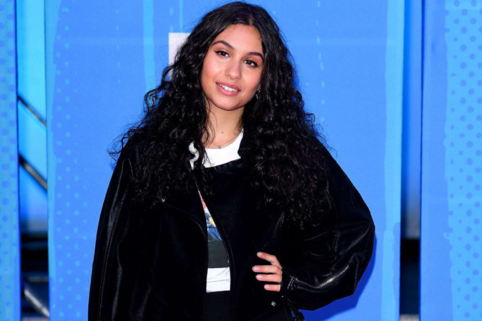Alessia Cara smiling with long curly hair, wearing a black jacket at an event with a blue background