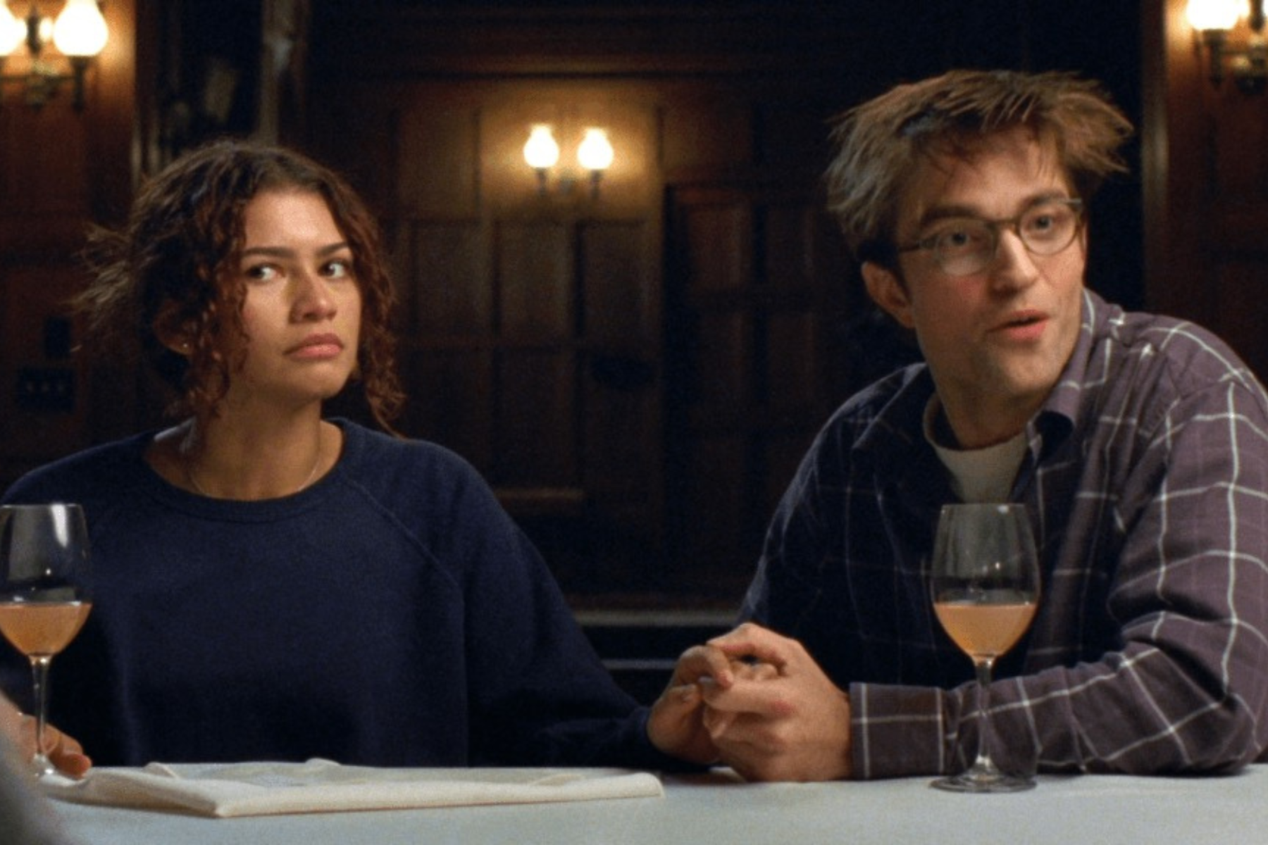Zendaya as Emma and Robert Pattinson as Charlie in The Drama. (Photo courtesy of A24). Zendaya and Robert Pattinson sit at a dinner table holding hands, reacting to someone offscreen