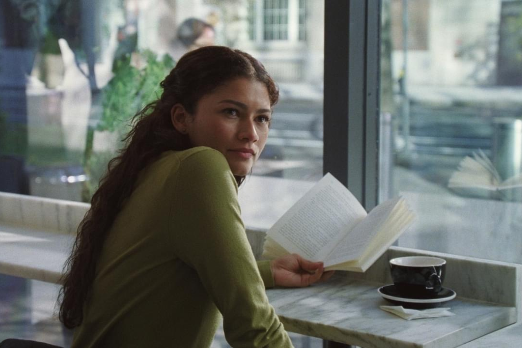 Zendaya as Emma in The Drama (Photo courtesy of A24). Zendaya sits by a window reading a book with a cup of coffee beside her