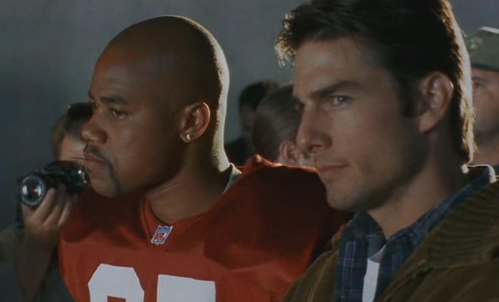 Jerry Maguire 30th Anniversary Review: Tom Cruise’s Most Heartfelt Work Still Holds Up Two men stand close together in a dim room: a bald man in a red NFL jersey and a serious-looking man in a plaid shirt, with a photographer in the background.