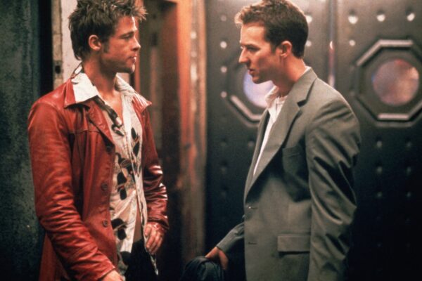Fight Club 4K Remaster Review: Tyler Durden Returns to the Big Screen in David Fincher’s Masterpiece Two men face each other in a dim hallway; one wears a red leather jacket over a patterned shirt, the other a gray suit and white shirt, holding a hat.