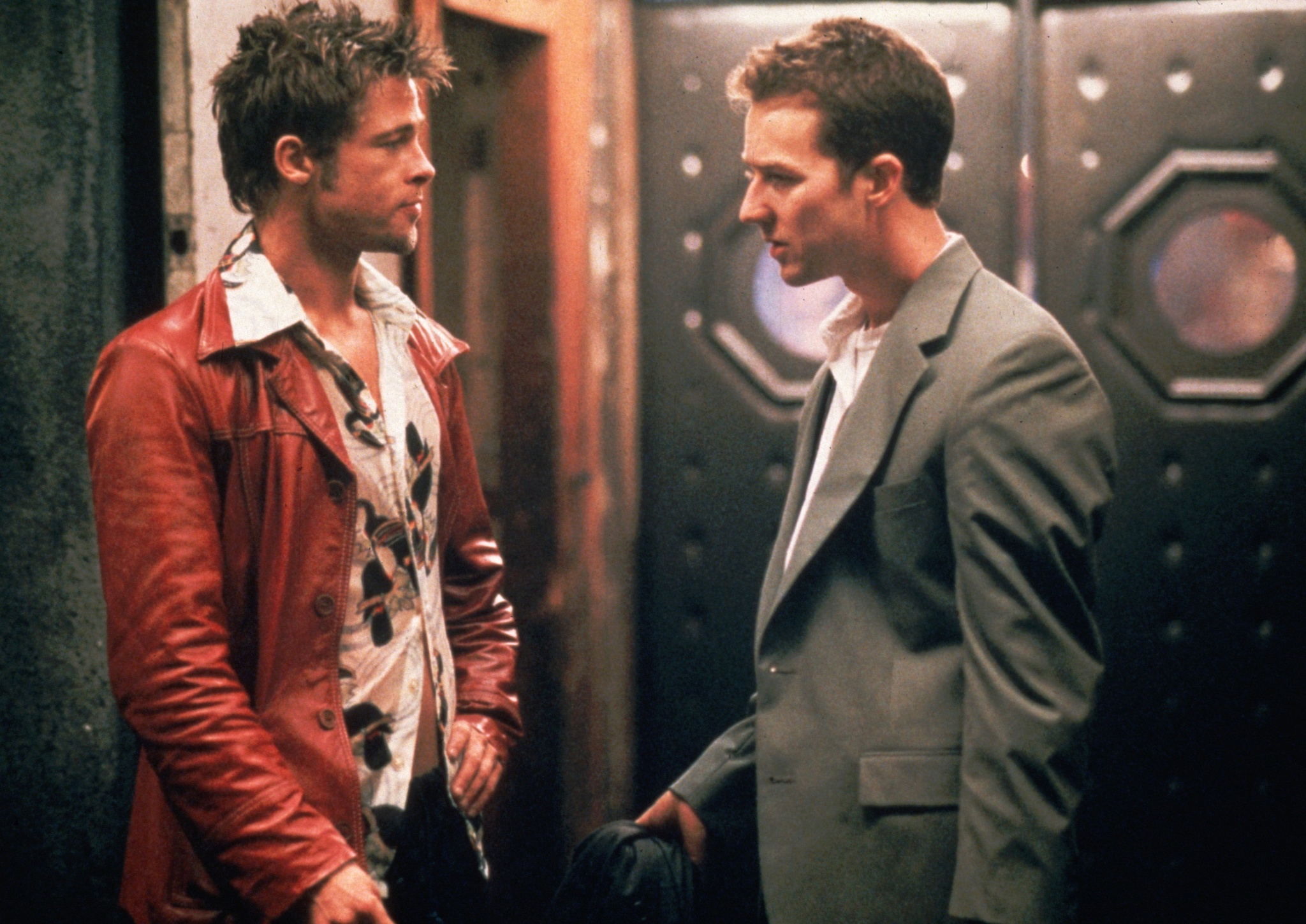 Fight Club 4K Remaster Review: Tyler Durden Returns to the Big Screen in David Fincher’s Masterpiece Two men face each other in a dim hallway; one wears a red leather jacket over a patterned shirt, the other a gray suit and white shirt, holding a hat.