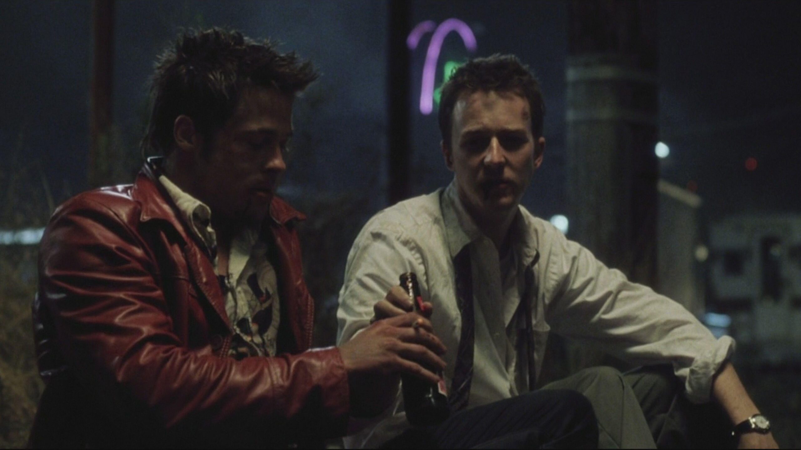 Fight Club 4K Remaster Review: Tyler Durden Returns to the Big Screen in David Fincher’s Masterpiece Two men sit together at night, each holding a bottle, one in a red leather jacket and the other in a light shirt, in a dim urban setting behind them.