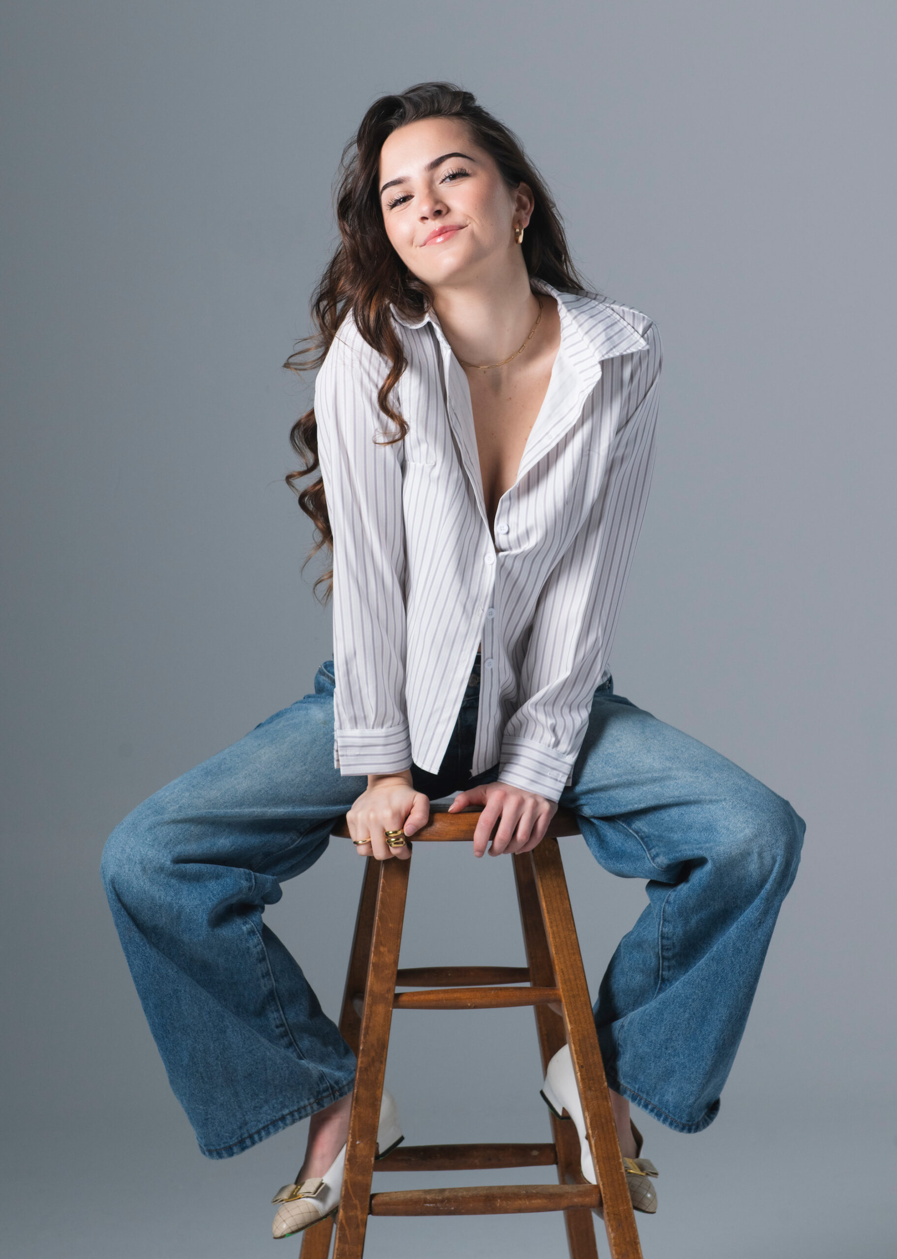 Hannah Cheramy Talks FROM Season 4 and Julie’s Journey Actress Hannah Cheramy, who stars in FROM Season 4, poses on a stool for a professional headshot in front of a gray backdrop.