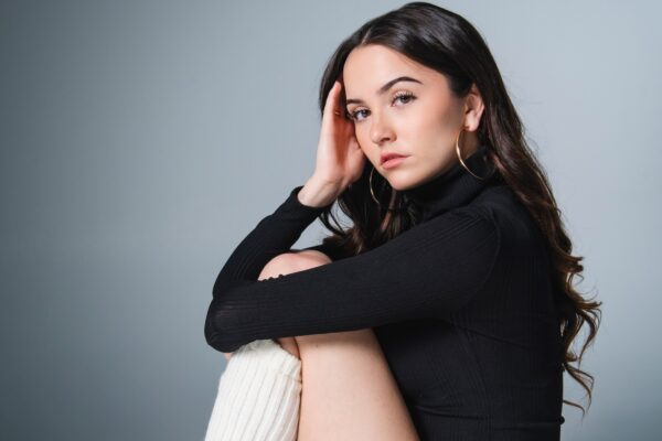 Actress Hannah Cheramy, who stars in FROM Season 4, poses for a professional headshot while wearing a black leotard with white leggings. She sits in front of a gray backdrop.