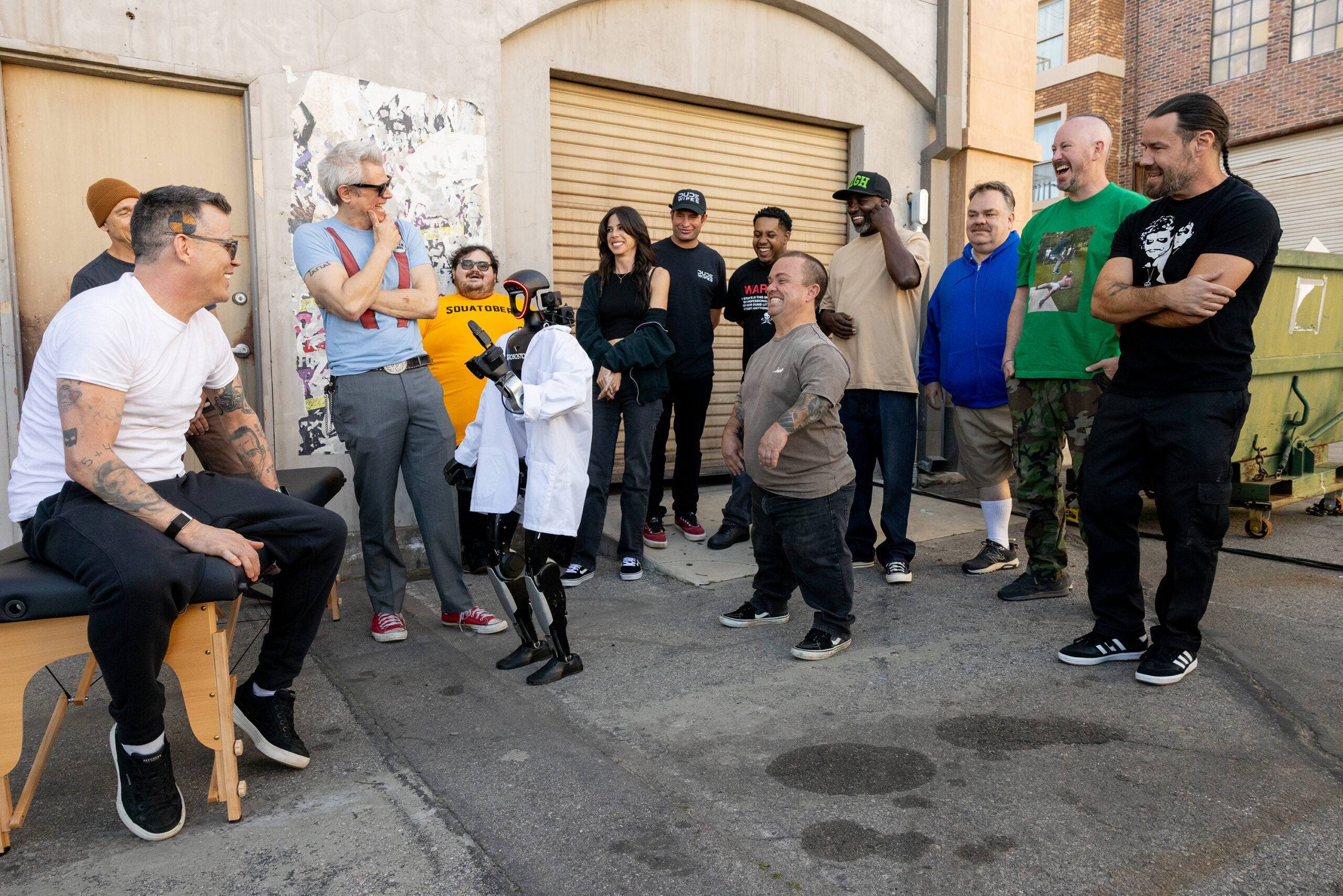 Jackass: Best and Last Trailer Dishes Out The Final, Most Painful Punishments to Celebrate A group of adults standing and smiling in an urban alleyway by a shuttered door and brick building.