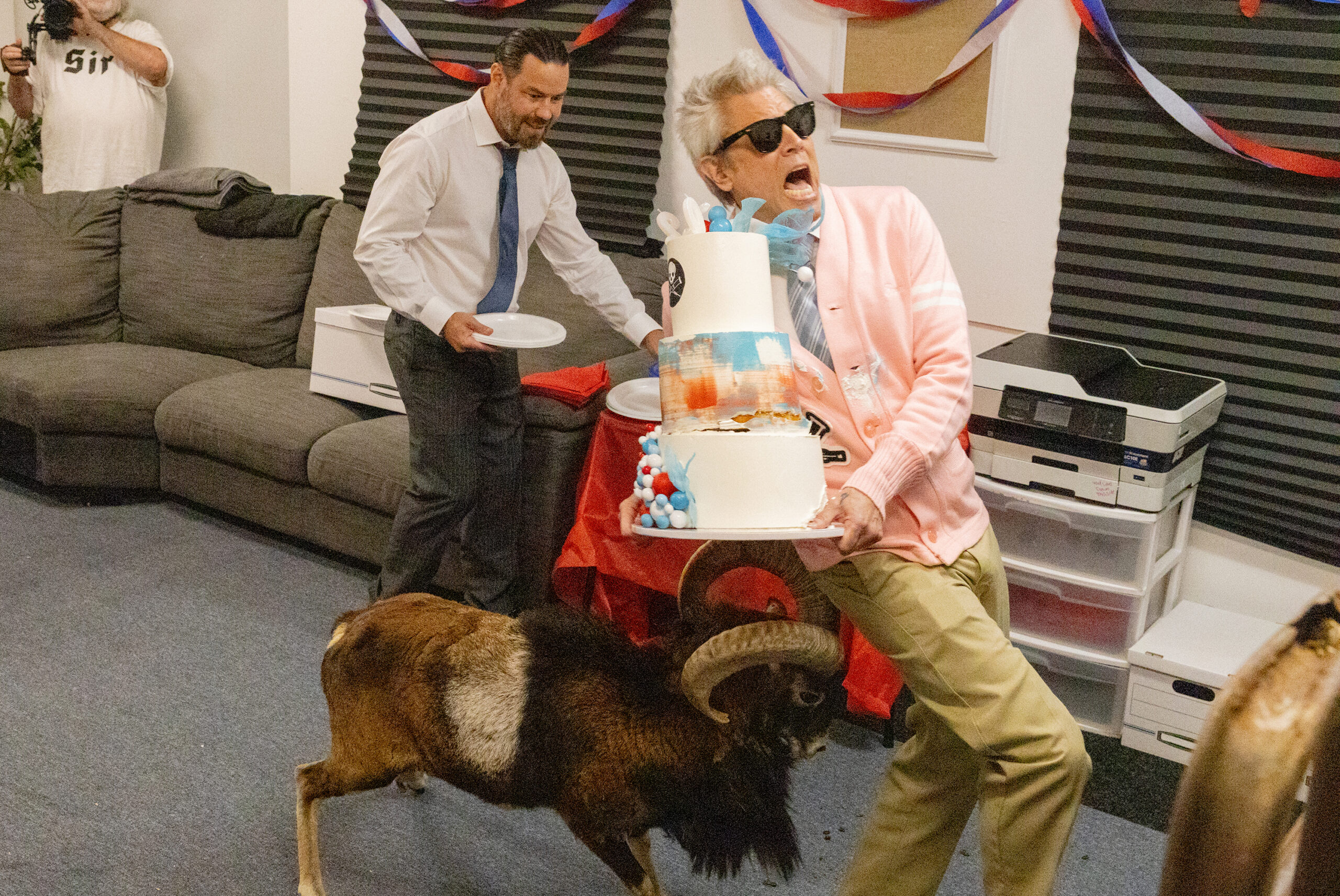 Jackass: Best and Last Trailer Dishes Out The Final, Most Painful Punishments to Celebrate A party scene with a two-tier cake and a person in a pink jacket wearing sunglasses while another man in a white shirt and blue tie serves cake; a large black furry mascot stands nearby as streamers hang in the room.