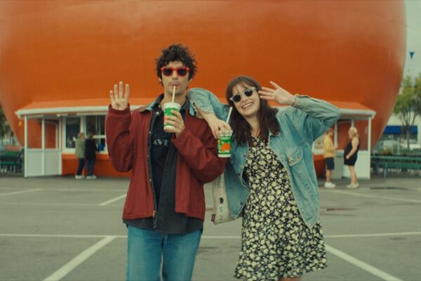 Mile End Kicks film still - Two friends pose for the camera with drinks in hand in front of a giant orange balloon at an outdoor fair, waving playfully.