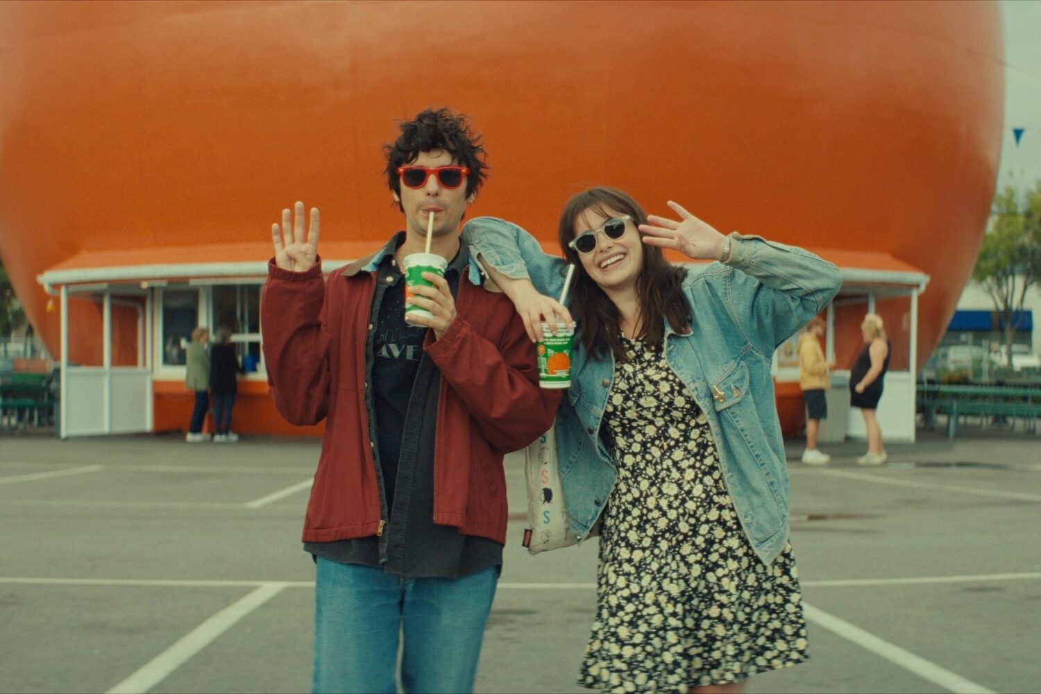 Mile End Kicks Review: Barbie Ferreira is a True Movie Star in a Finely Made Coming-of-Age Story Mile End Kicks film still - Two friends pose for the camera with drinks in hand in front of a giant orange balloon at an outdoor fair, waving playfully.