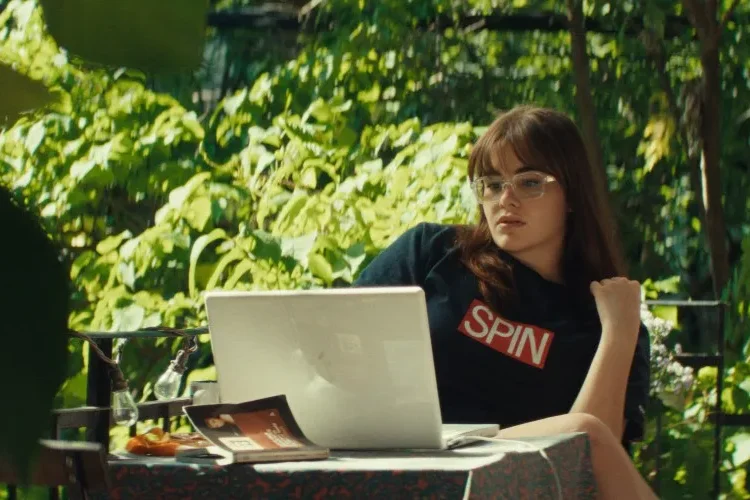 Mile End Kicks Review: Barbie Ferreira is a True Movie Star in a Finely Made Coming-of-Age Story Woman with glasses sits at an outdoor table using a laptop, surrounded by lush green plants.
