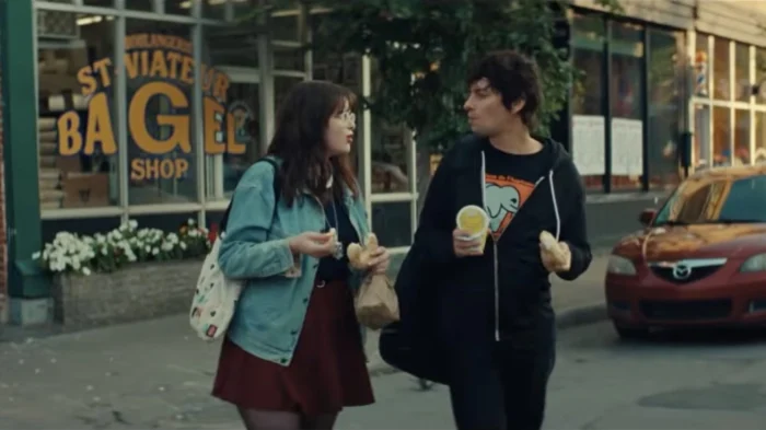 Mile End Kicks Review: Barbie Ferreira is a True Movie Star in a Finely Made Coming-of-Age Story Two people walking on a city sidewalk, eating bagels and chatting, with a bakery storefront in the background.