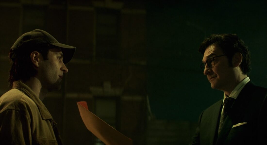 Daredevil: Born Again Season 2 Episode 5 Review: ‘The Grand Design’ Is A Greater Metaphor Two men face each other in a dimly lit room; one wears a cap and jacket, the other a suit holding an orange folder.