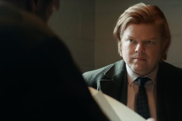 Man in a suit and tie with blond hair, looking serious while examining a document in a dimly lit room