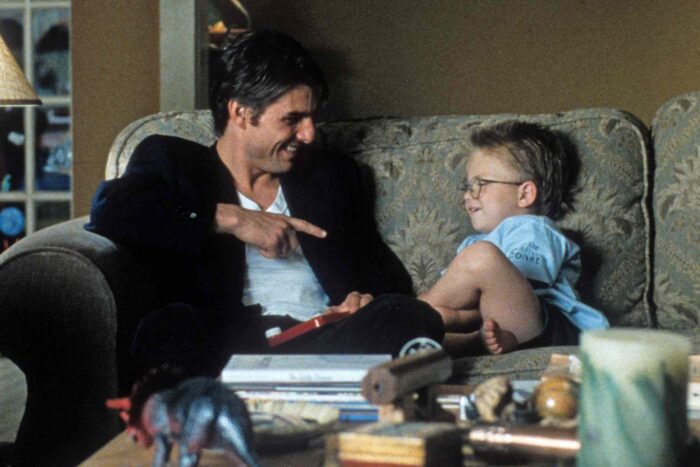 Jerry Maguire 30th Anniversary Review: Tom Cruise’s Most Heartfelt Work Still Holds Up Father and young son sit on a patterned sofa, smiling as they share a playful moment and point at each other.
