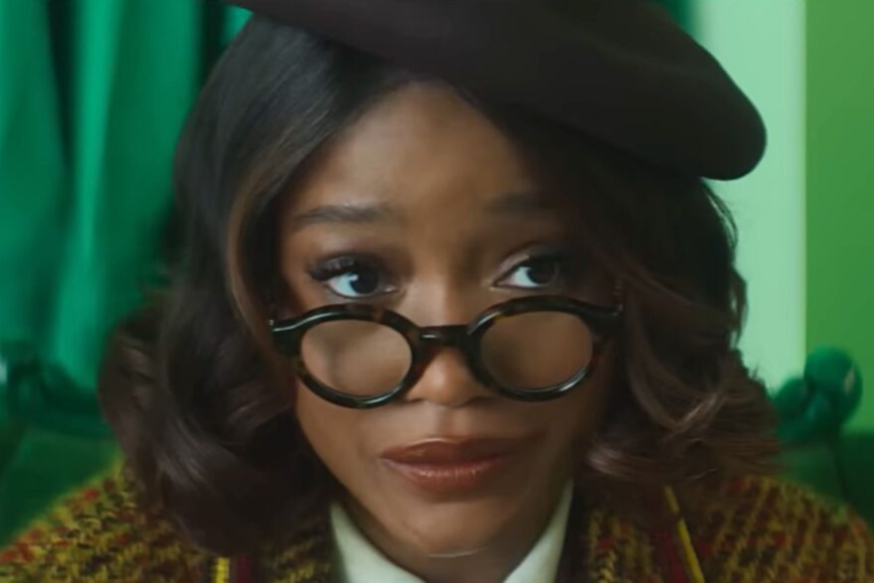 I Love Boosters Review: Stylish, Strange, and Slightly Uneven Keke Palmer, wearing round glasses and a black beret, looks over the rim of her glasses while seated indoors.