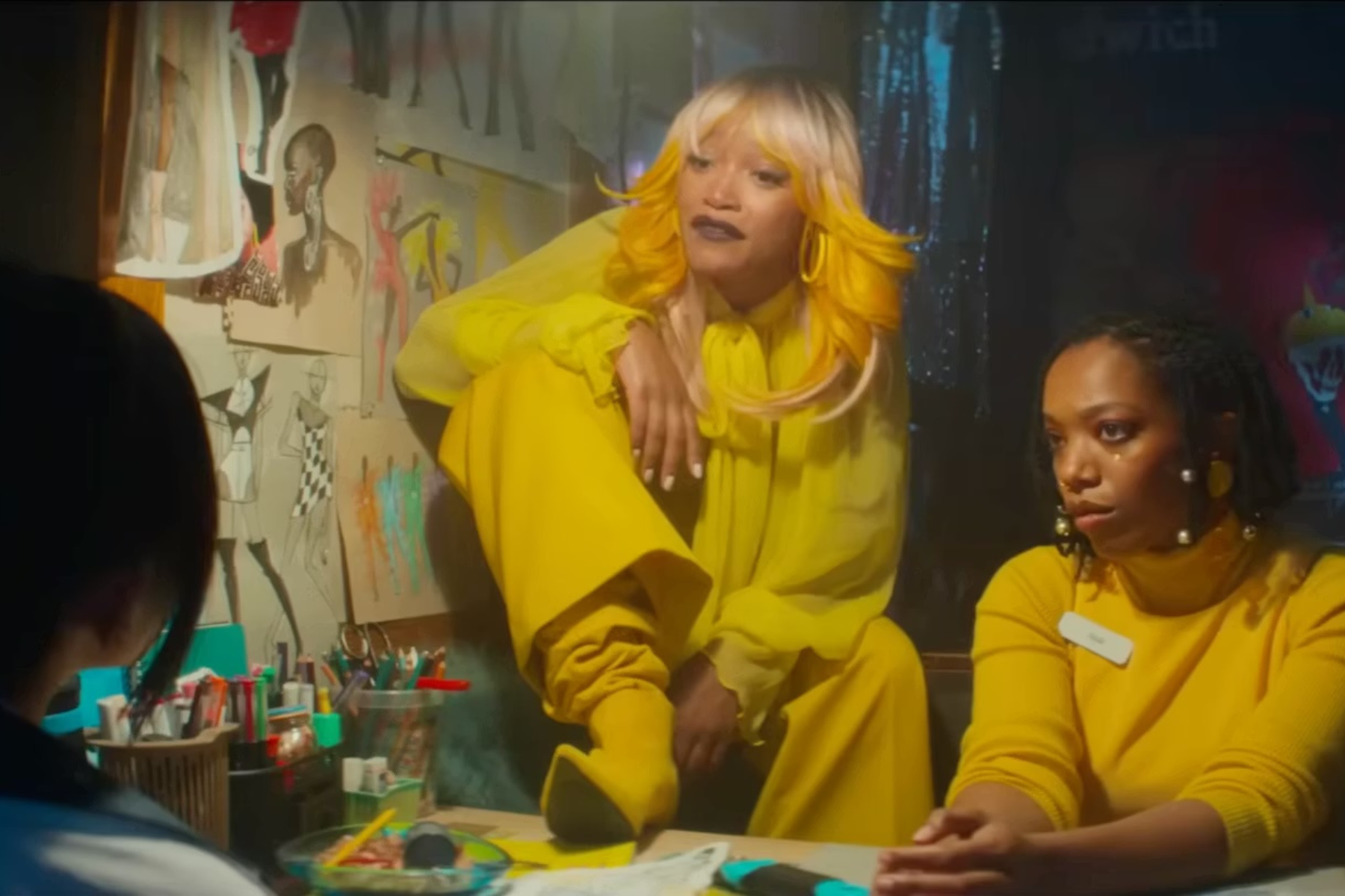 I Love Boosters Review: Stylish, Strange, and Slightly Uneven Keke Palmer and Naomi Ackie sit at a cluttered table while discussing plans in a dimly lit room filled with fashion sketches and supplies.