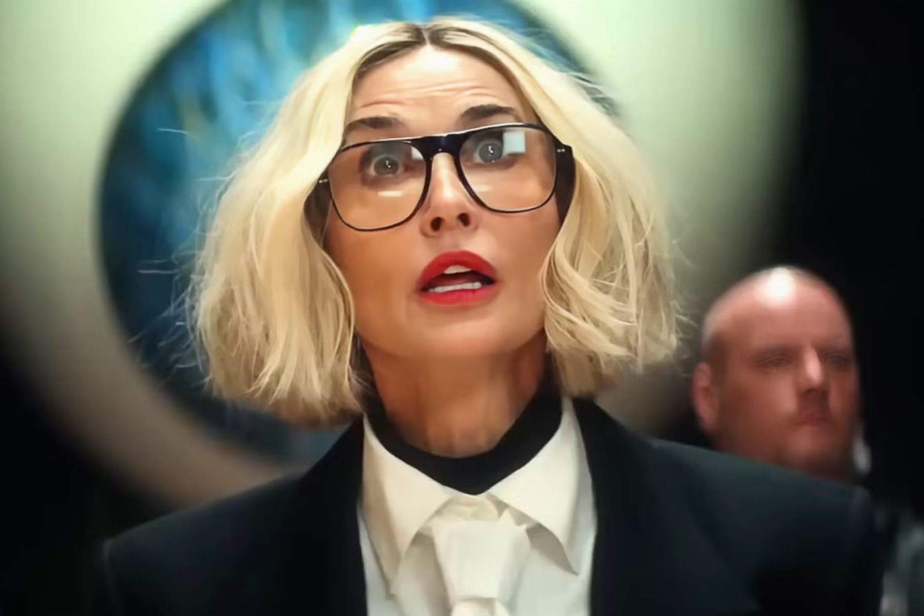 I Love Boosters Review: Stylish, Strange, and Slightly Uneven Demi Moore, wearing oversized glasses and a black-and-white suit, reacts with surprise while looking through a circular opening.