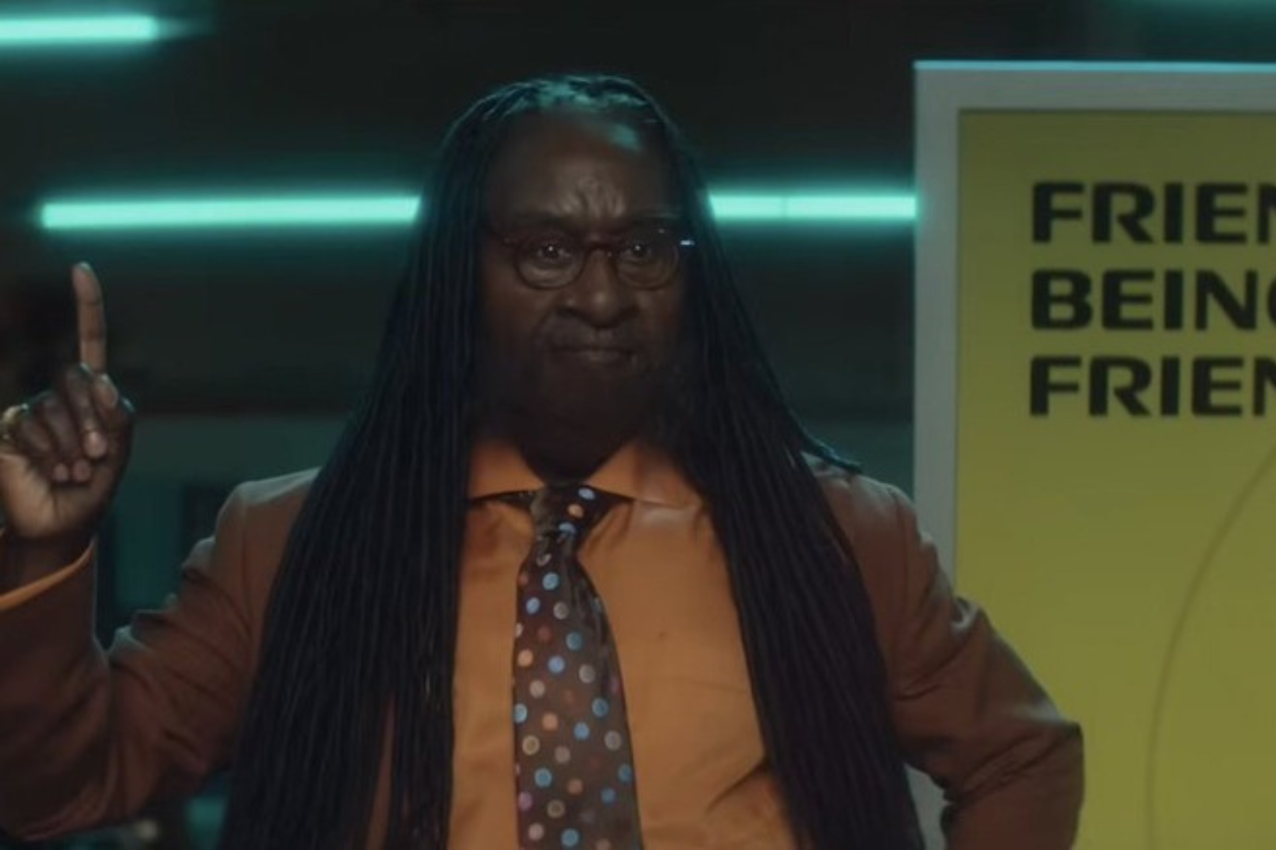 I Love Boosters Review: Stylish, Strange, and Slightly Uneven Don Cheadle gestures with one finger raised while speaking during a presentation in a dimly lit room.