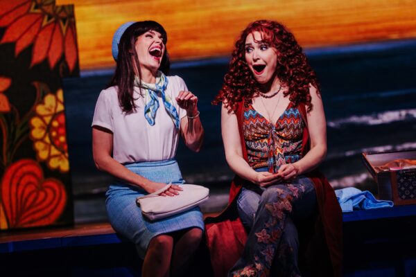 Two women on a colorful stage share a laugh during a performance, seated side by side.