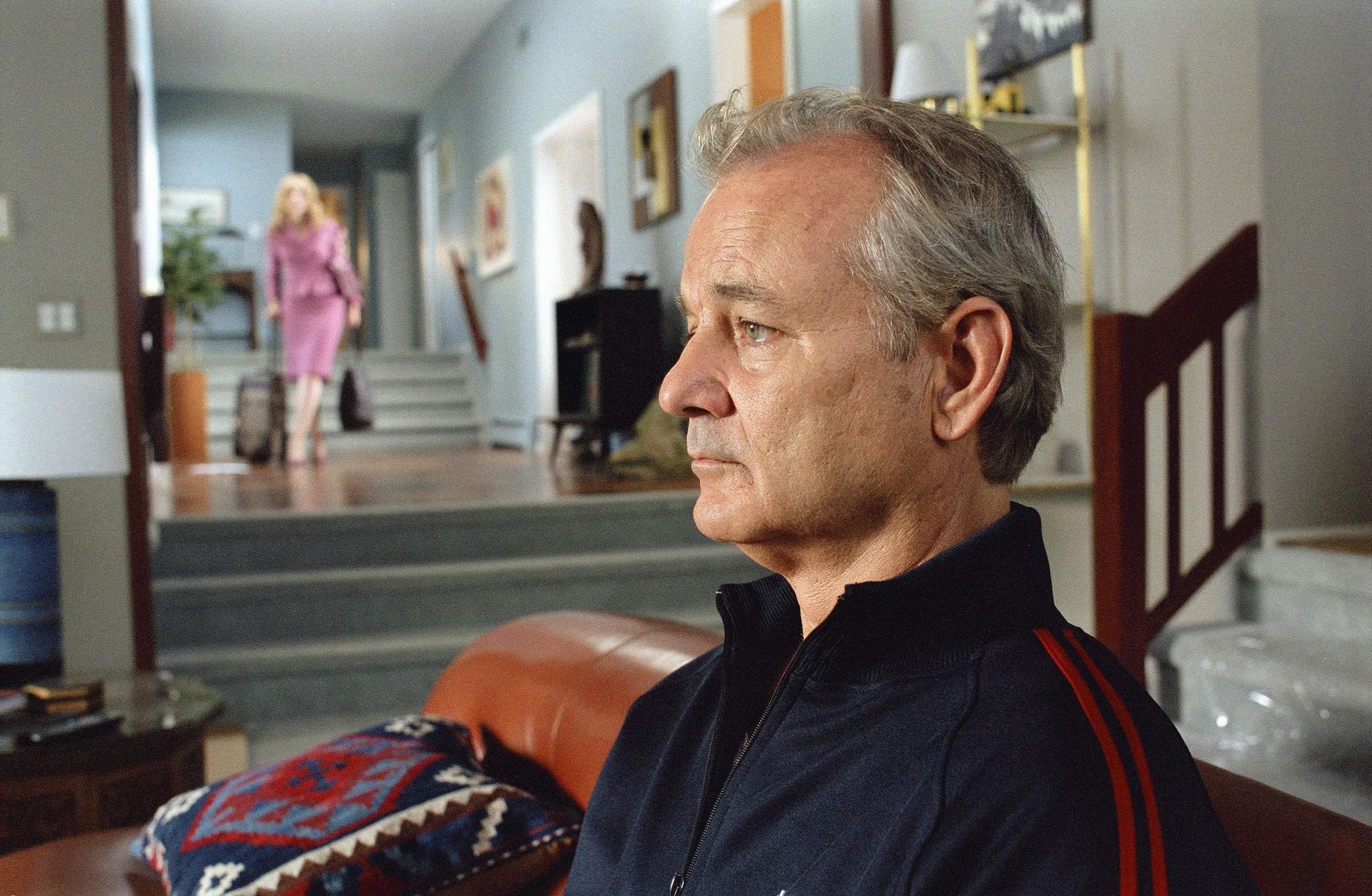 The 12 Greatest Movies Featuring Bill Murray Close-up side profile of an older man in a living room, with a woman in pink walking in the background near the stairs.