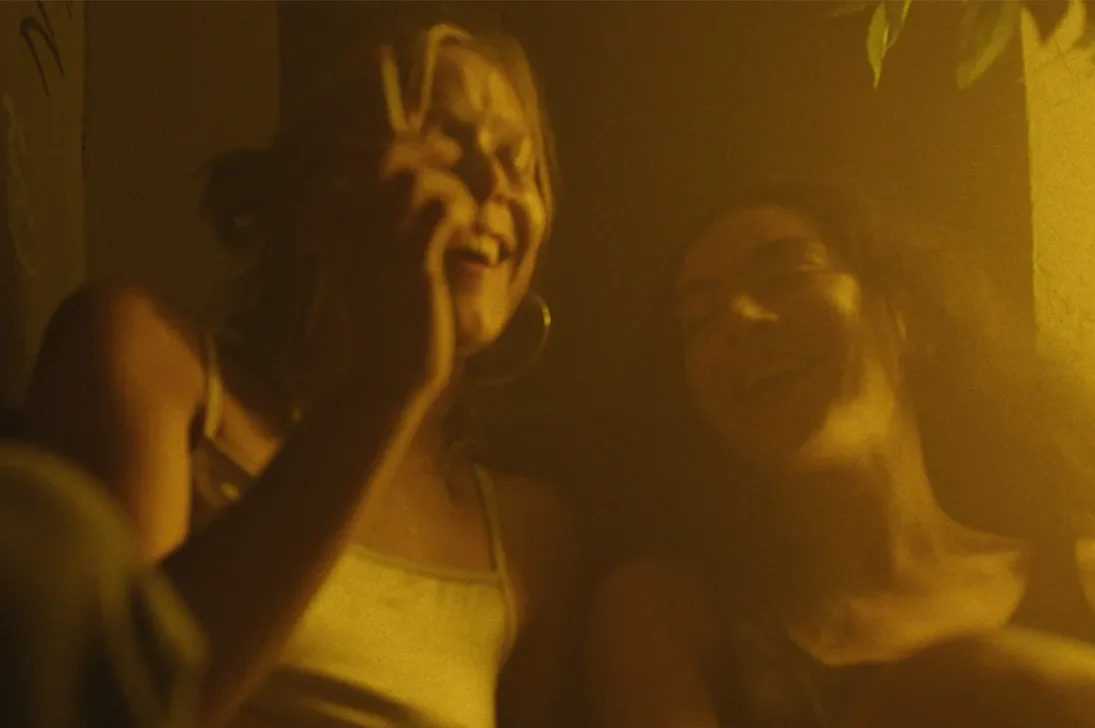 Erupcja Review: Charli XCX Stars in a Thought-Provoking Film for the Art House Crowd Two women smiling at the camera in a warm, yellow-lit indoor setting, likely at a social gathering.