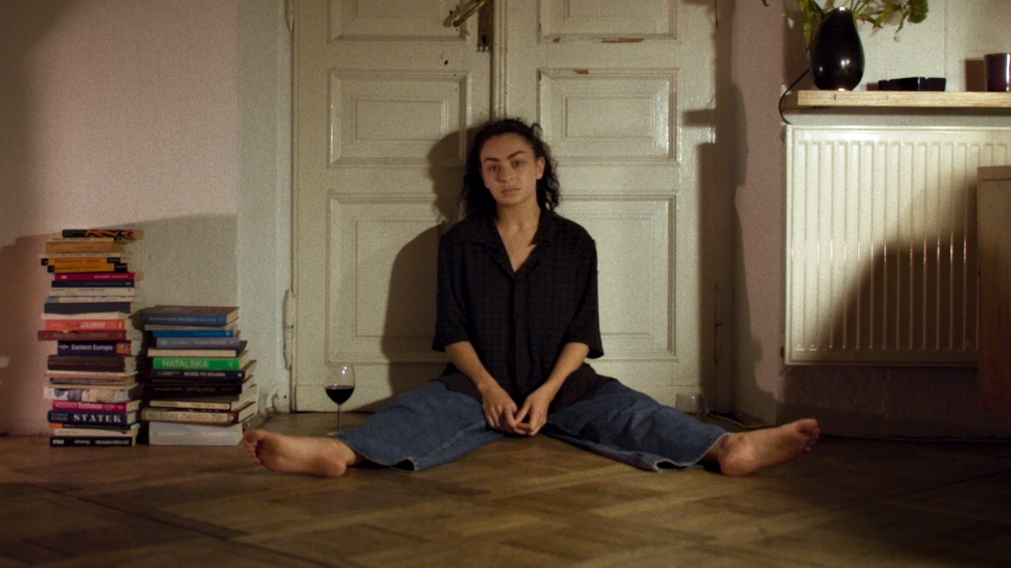 Erupcja Review: Charli XCX Stars in a Thought-Provoking Film for the Art House Crowd Person sits cross‑legged on a wooden floor, leaning against a pale paneled door, with a tall stack of books on the left and a glass of red wine nearby.