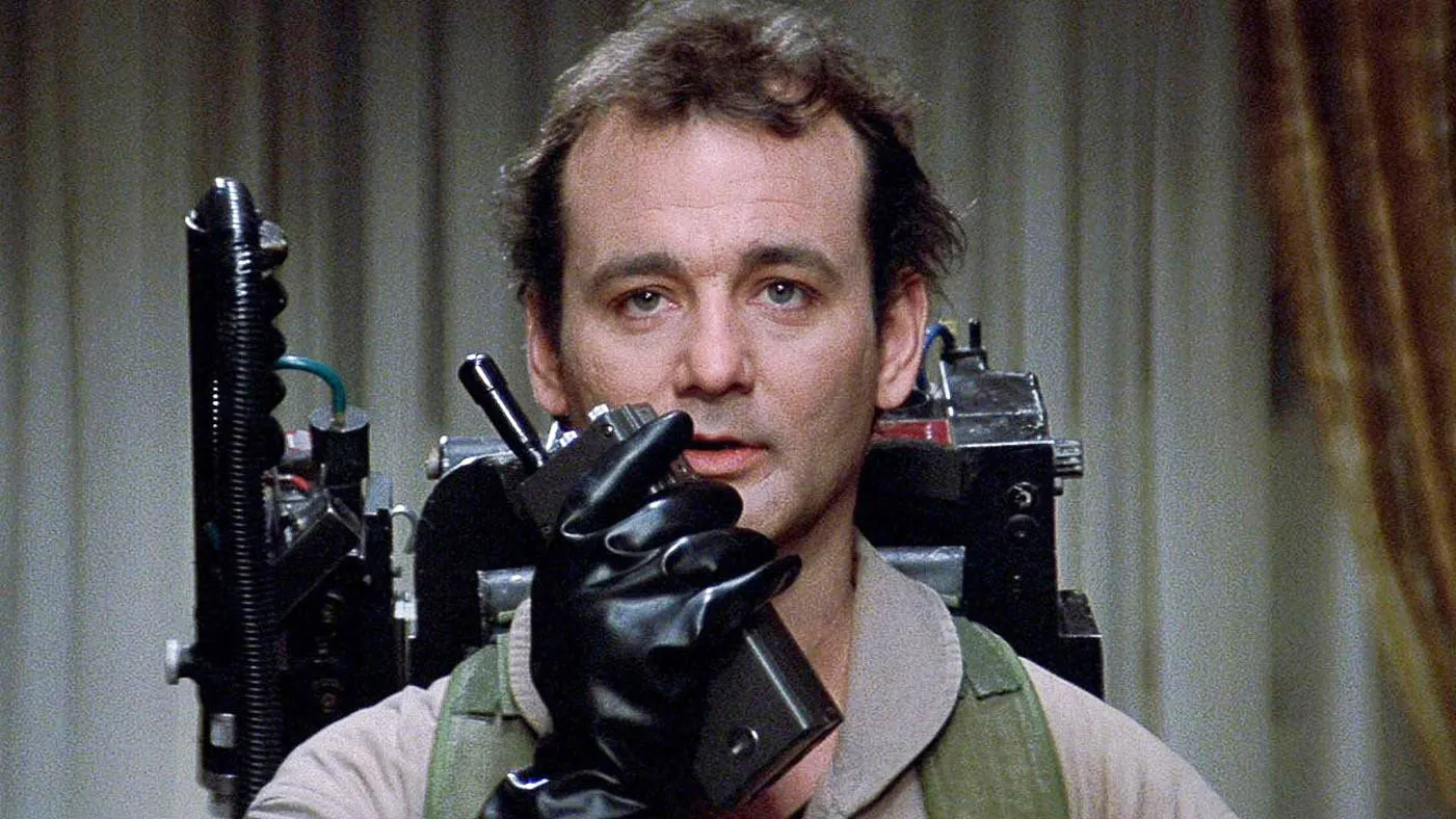 The 12 Greatest Movies Featuring Bill Murray Man in a beige jumpsuit with a proton-pack style backpack, wearing a black glove and holding a handheld device toward the camera.