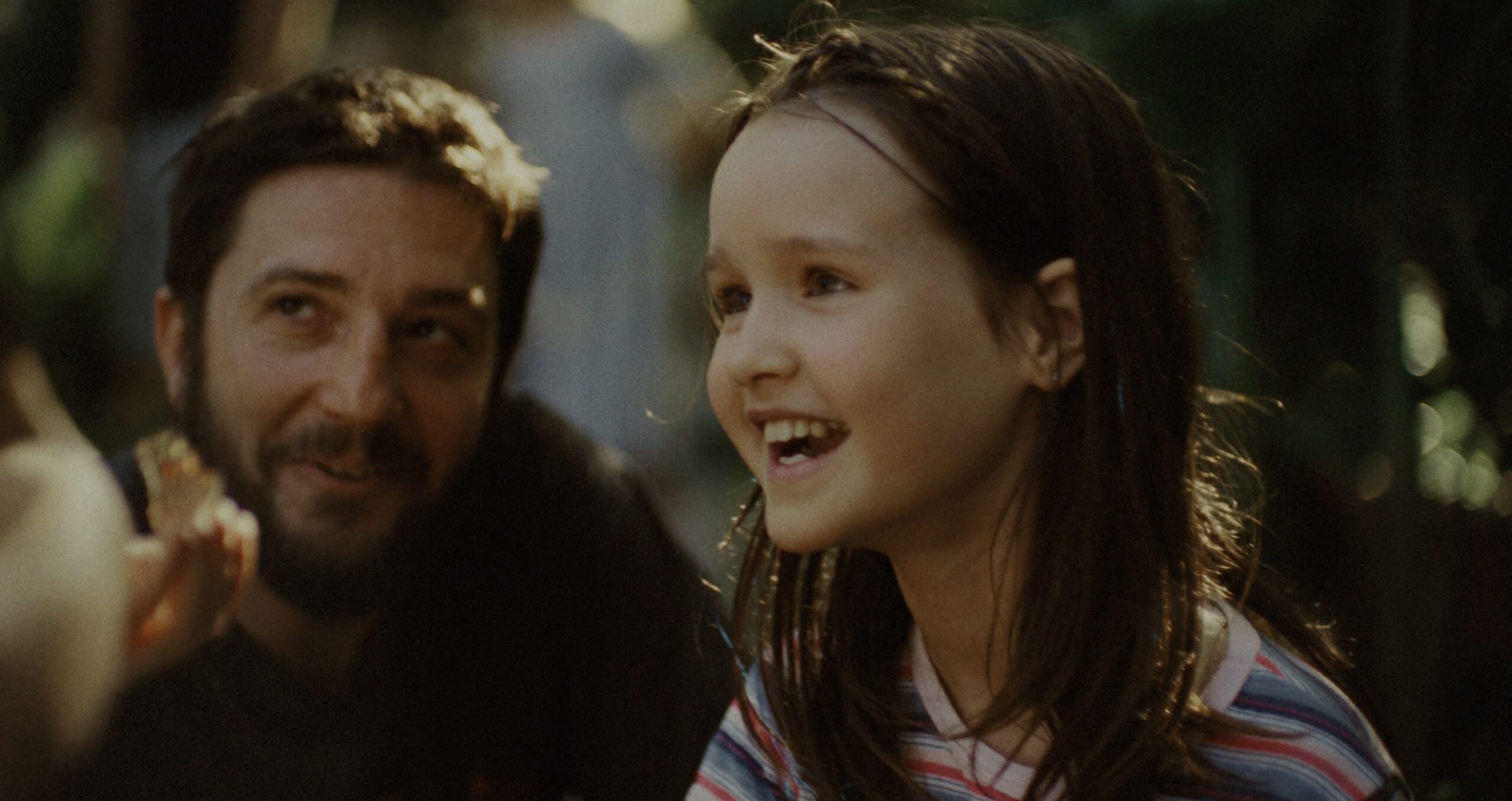 Omaha Review: John Magaro is Amazing in a Dramatic Film That Packs a Wallop Girl with long brown hair laughing outdoors, with a bearded man nearby in soft focus.