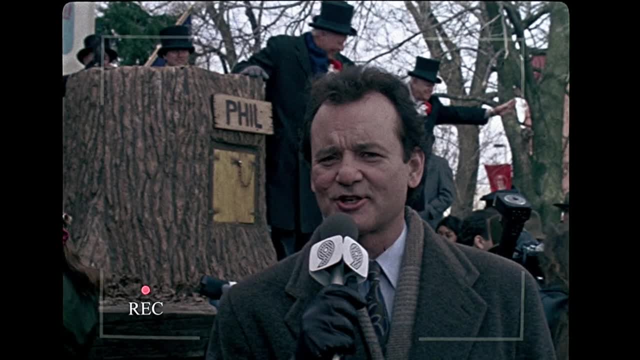 The 12 Greatest Movies Featuring Bill Murray News reporter speaks into a microphone at an outdoor event; a coffin-like prop labeled PHIL sits behind him with men in top hats in the background.