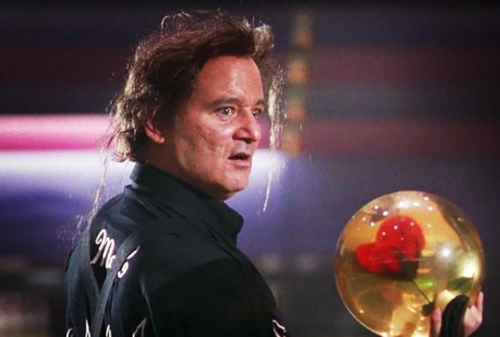The 12 Greatest Movies Featuring Bill Murray Man in a dark jacket looks surprised while holding a reflective gold trophy with a red center at a nighttime event.