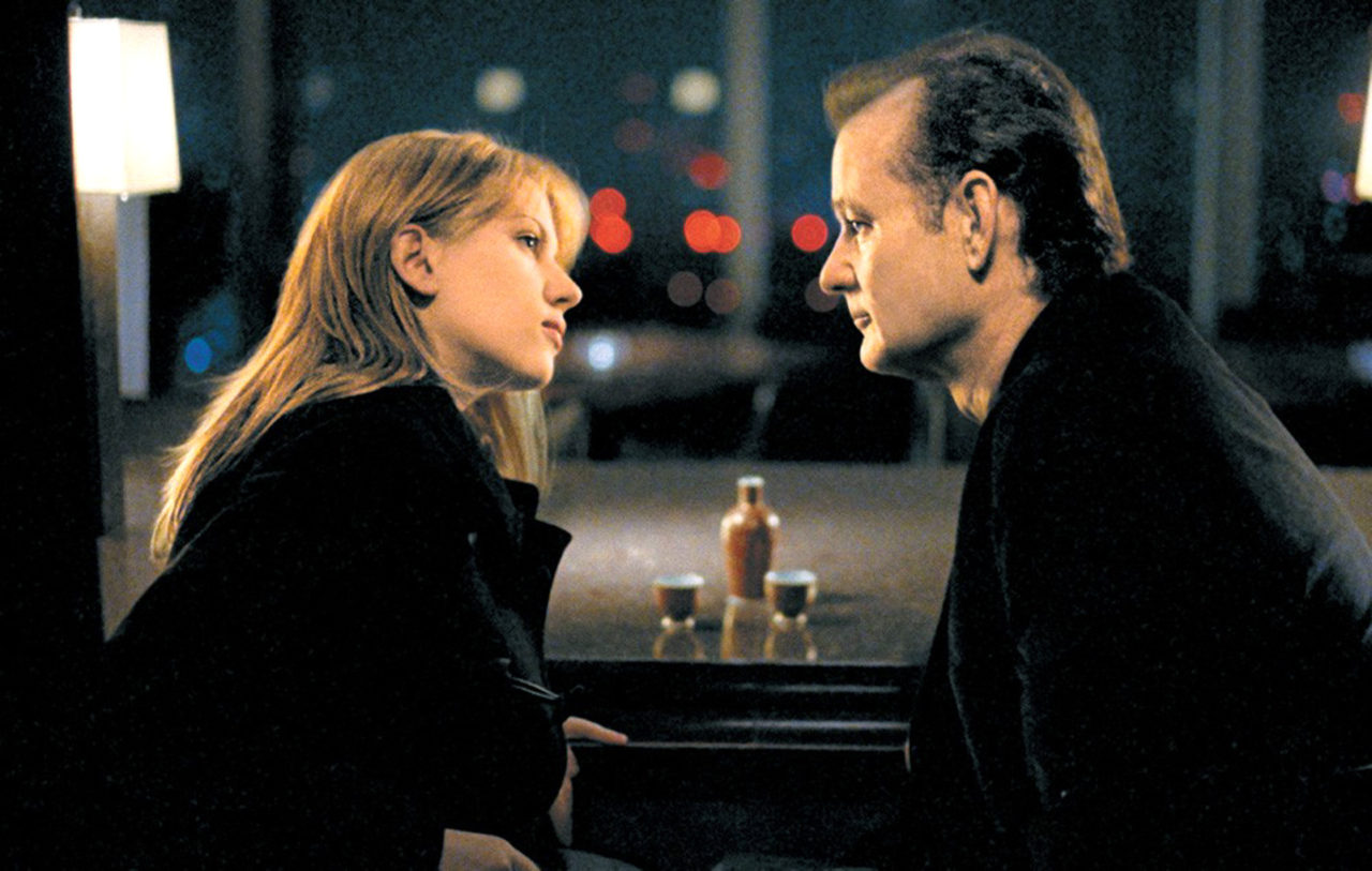 The 12 Greatest Movies Featuring Bill Murray Two people sit facing each other at a windowed table with a bottle and two glasses, nighttime city lights in the background, suggesting an intimate conversation.