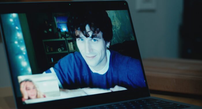 Maximum_Pleasure_Guaranteed_Photo_010102 - Eulalie Magazine Maximum Pleasure Guaranteed Season 1 Episode 1 and 2 Review: Magnets / YABA Man with dark hair and blue shirt on a laptop video call, blue-eyed, in a dimly lit room; reflected image on screen