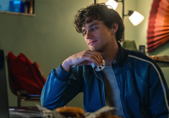 Maximum_Pleasure_Guaranteed_Photo_010104 - Eulalie Magazine Maximum Pleasure Guaranteed Season 1 Episode 1 and 2 Review: Magnets / YABA Young man with curly hair in a blue jacket sits at a table, looking thoughtful while holding a small object near his mouth in a cozy room.