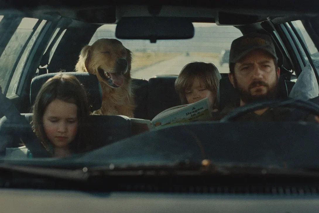 Omaha Review: John Magaro is Amazing in a Dramatic Film That Packs a Wallop Man drives a car with two kids in the cabin and a golden retriever in the back seat; one child reads a book.