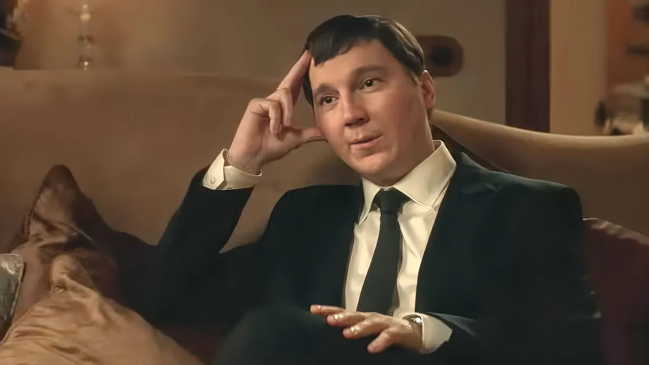 The Wizard of the Kremlin Review: Paul Dano is Brilliant in This Chilling Film Man in a dark suit sits on a sofa, one hand at his temple in a thoughtful pose inside a warmly lit room.