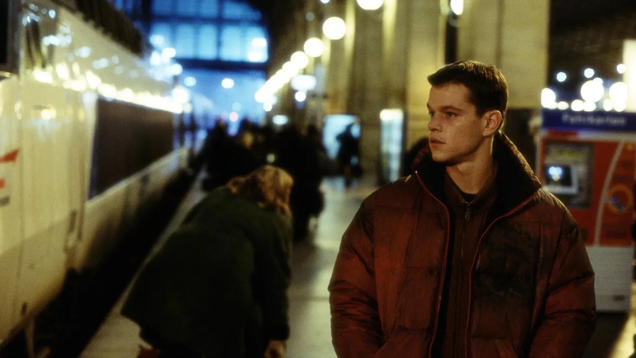 The 12 Best Movies Featuring Matt Damon in a Principal Role Young man in a brown puffer jacket waiting on a train platform at night, with a train to the left and blurred passengers in the background.