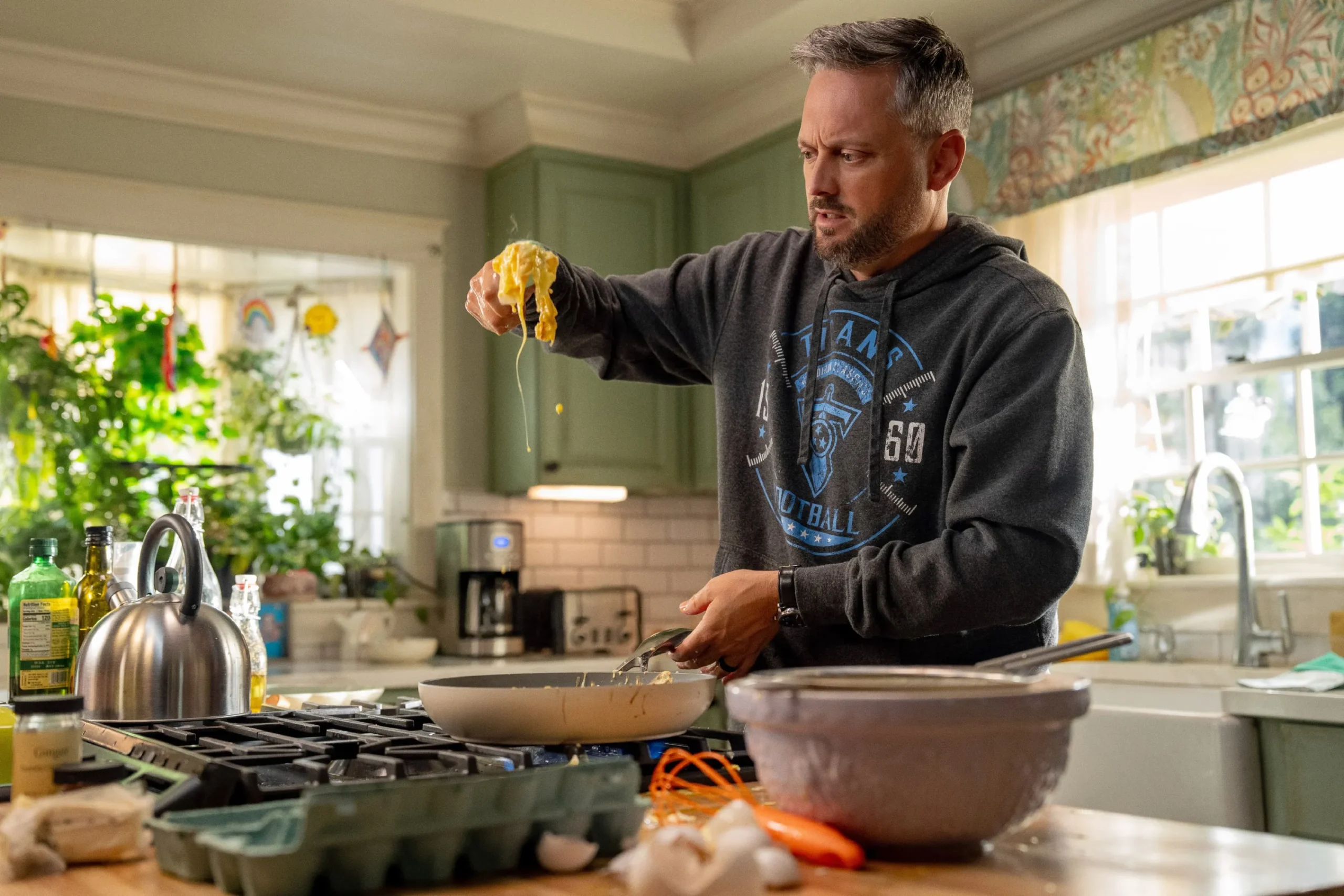 The Breadwinner (Photo courtesy of Sony Pictures) - Eulalie Magazine The Breadwinner Review: A Mr. Mom-Type Comedy that Loses Its Way Despite Funny Scenes Man in a dark hoodie stands at a kitchen island, lifting a strand of melted food from a frying pan with a spatula.