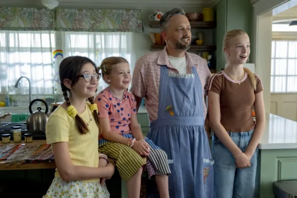 Father in a blue apron stands with three smiling kids in a bright, cozy kitchen.
