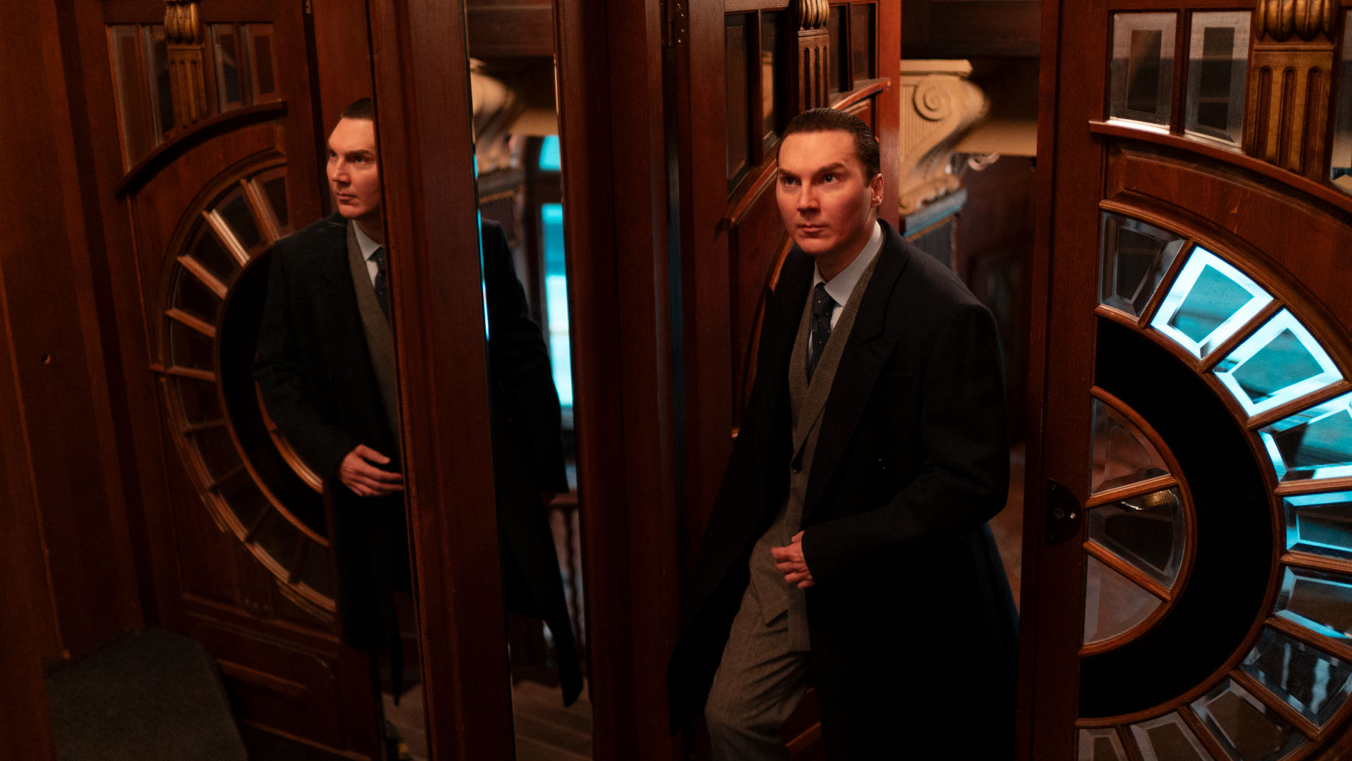 The Wizard of the Kremlin Review: Paul Dano is Brilliant in This Chilling Film Man in a suit and vest steps through a circular, wood‑paneled revolving door with glass panels in a dimly lit, vintage setting at interior.