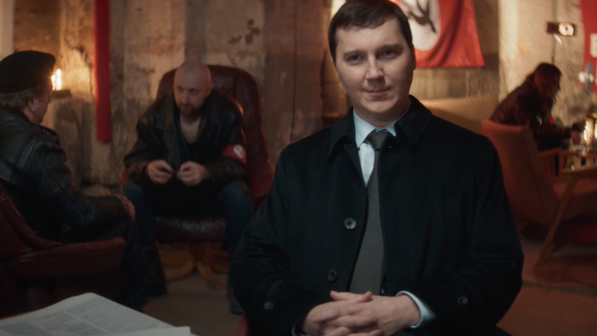 The Wizard of the Kremlin Review: Paul Dano is Brilliant in This Chilling Film Foreground: man in a suit with hands folded sits at a table; blurred background features two men and a red banner in a rustic, dim room.
