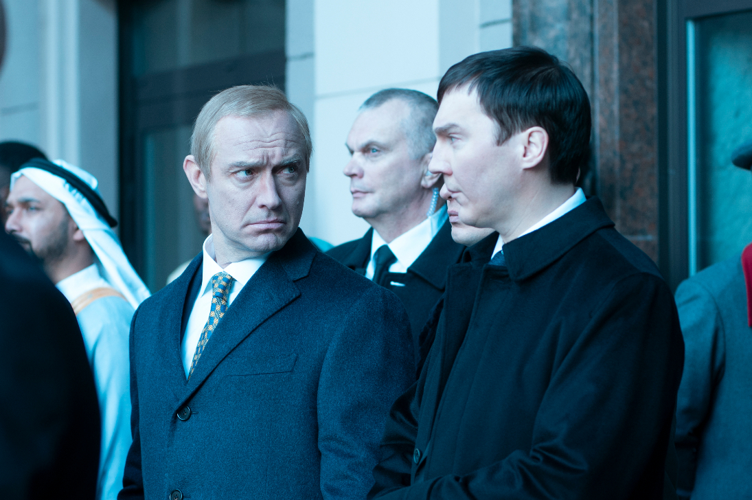 The Wizard of the Kremlin Review: Paul Dano is Brilliant in This Chilling Film Two men in coats stand outside a building; the man in the gray suit with a patterned tie eyes the younger man in black with a stern expression, as others look on.