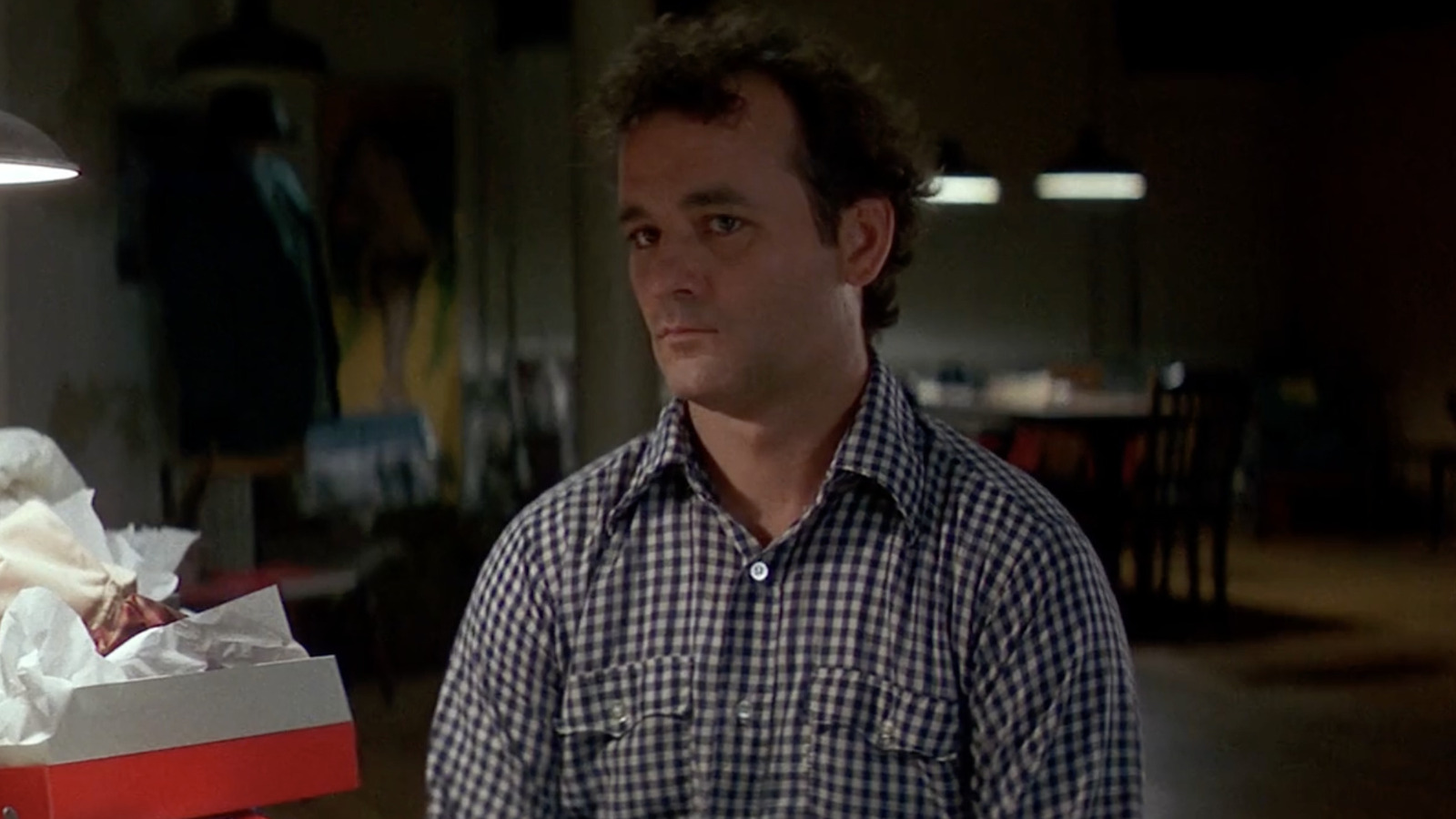 The 12 Greatest Movies Featuring Bill Murray Man in a blue-and-white plaid shirt stands in a dim room, looking off to the left, with a wrapped gift box in the foreground.