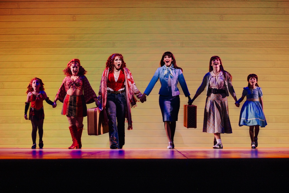 Beaches: A New Musical Review: A Beautiful Tale of Friendship Told in a Heartwarming Fashion Six performers in colorful vintage costumes stand in a line on a stage, holding hands against a yellow backdrop.