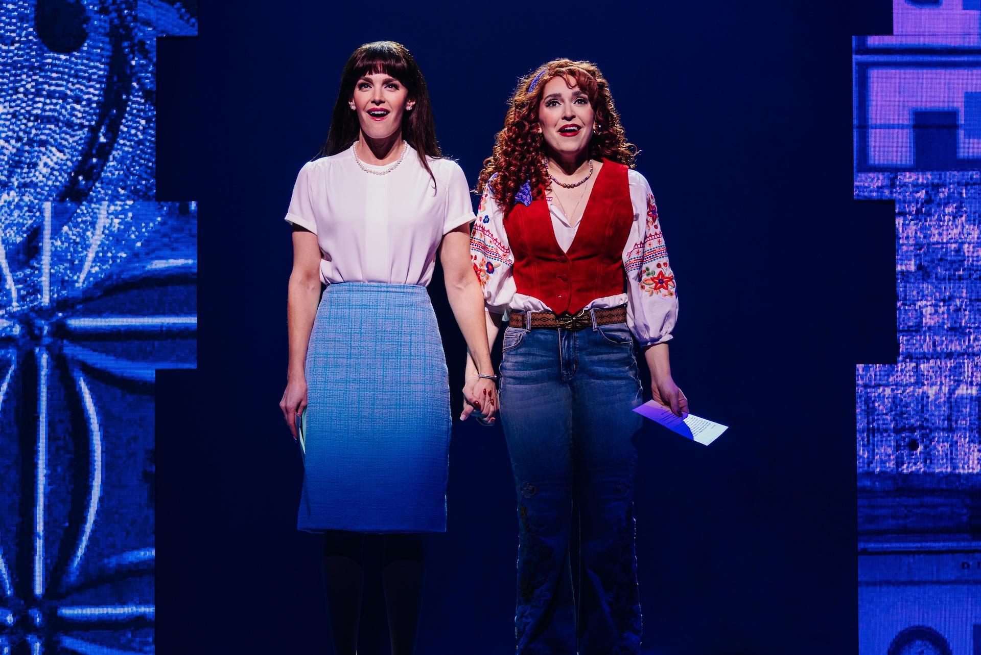 Beaches: A New Musical Review: A Beautiful Tale of Friendship Told in a Heartwarming Fashion Two women stand on a blue-lit stage, holding hands and smiling; one wears a white blouse with a blue skirt, the other a floral blouse with a red vest and jeans.