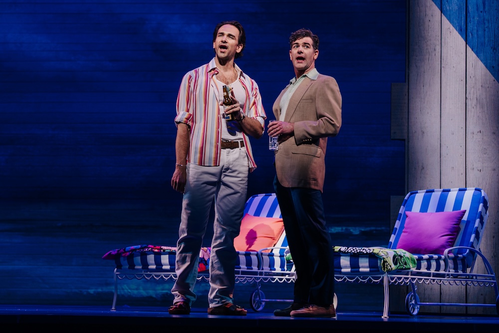 Beaches: A New Musical Review: A Beautiful Tale of Friendship Told in a Heartwarming Fashion Two men stand on a stage holding bottles, beside striped beach chairs with colorful cushions and blankets in a theatrical set.