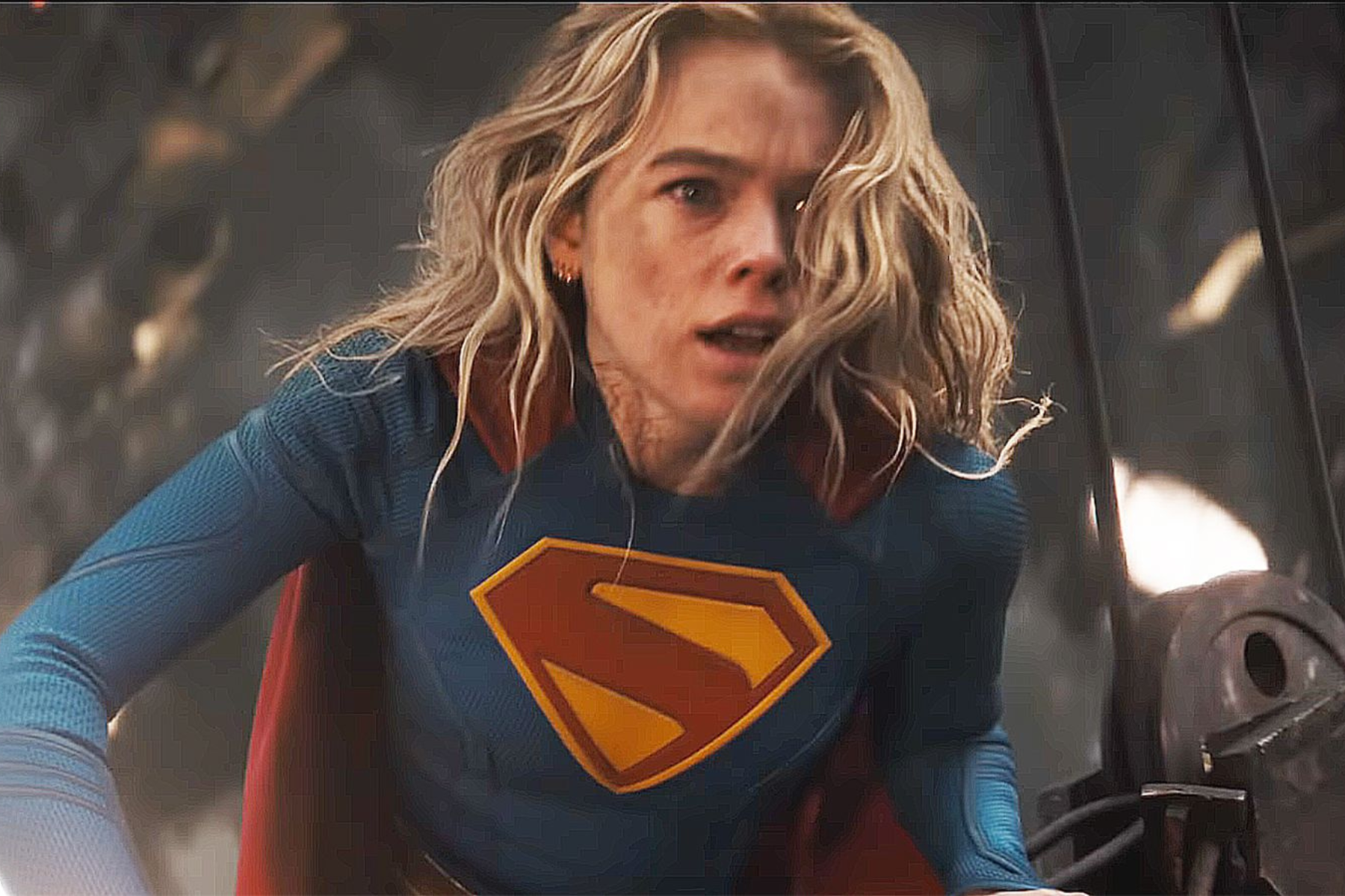 Supergirl Review: Strong Performances Can’t Save an Uneven Adventure Female superhero in a blue suit with a yellow shield emblem on her chest, leaning forward in an action scene.